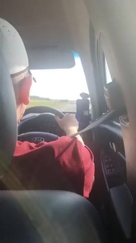 This is a video capturing a journey from the interior of a car. The scene is a roadside view from the passenger seat, looking out the side window and forward towards the driver. The driver, wearing a red shirt, is visible from behind, holding the steering wheel of a Kia. A red cloth item, possibly a bag or blanket, rests on the passenger seat. The road appears to be a highway with a metal guardrail on the right side, bordered by fields. The sky is bright with a strong sun, suggesting it is daytime, likely a sunny day. The overall mood seems relaxed, consistent with a road trip. No text is clearly visible other than the "KIA" logo on the steering wheel. The location is identified as Stonehaven, United Kingdom.