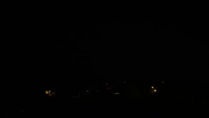 The video captures a brief, faint flash of lightning in the distance over a dark, silhouetted landscape. The landscape appears to be a rural or suburban area with a few scattered lights from buildings visible on the horizon. The sky is completely black, indicating nighttime, and no other atmospheric conditions like rain are discernible. The lightning flash is quick and subtle, barely illuminating the scene before returning to darkness. There are no people, vehicles, or discernible objects in the foreground or mid-ground of the shot. The overall impression is one of a distant storm observed from afar.