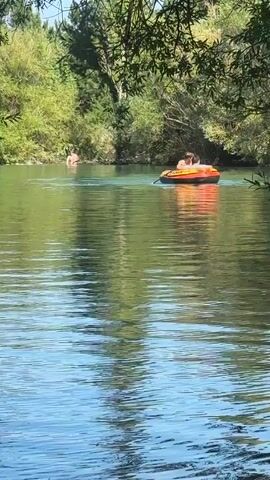 The video shows a wide, calm river on a sunny day. Two people are in an orange inflatable raft, with one person paddling. In the background, on the far bank of the river, at least two other people are visible in the shallow water near the shore. Dense green foliage and trees line the banks of the river, with sunlight dappling through the leaves and reflecting on the water's surface. The overall scene depicts a peaceful outdoor recreational setting.