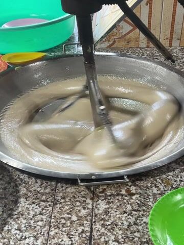 A large metal mixer in a kitchen mixes a beige mixture in a large stainless steel bowl. The mixer is stationary, and the beaters rotate in the mixture. Around the bowl are several plastic bowls in colors of green, yellow, and pink. The bowl sits on a mottled brown countertop.