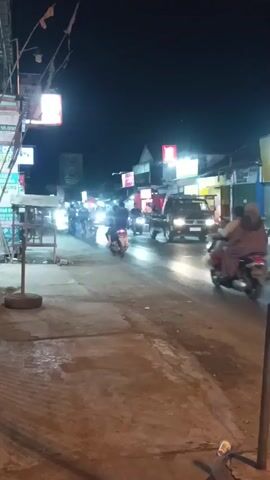 The video shows a street scene at night. Several motorcycles and a car are visible on the road, moving from left to right. The street is lined with buildings that have illuminated signs, suggesting they are shops or businesses. The pavement on the left side of the street is visible, with a tire leaning against a pole. The overall atmosphere is that of a busy street at night.