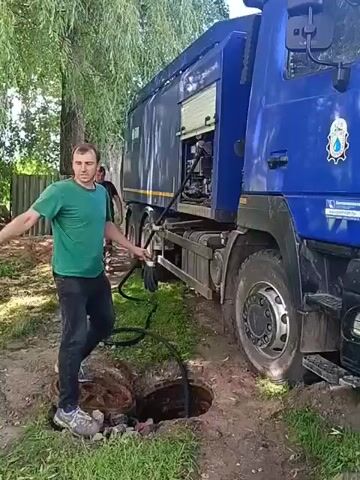 The video shows two men near a large blue truck that appears to be a vacuum truck, possibly for sewage or waste removal. The truck is parked on a dirt path next to a grassy area with trees. In the foreground, there is an open manhole with a black hose extending from the truck and into the manhole. The man on the left, wearing a green t-shirt and dark pants, is holding a black hose and looking towards the truck. The man on the right, wearing a dark work outfit with reflective stripes on his pants, is standing over the manhole, possibly preparing to work in it or manage the hose. The scene suggests a municipal service being performed, likely cleaning or emptying a sewer or septic tank. The lighting suggests it is daytime, and the weather appears to be fair. There is some text visible on the side of the truck, including a logo that seems to depict a water droplet. The location is identified as Nesvizh, Belarus.