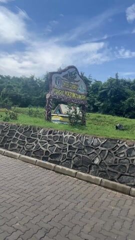 The video shows a sign that reads "Ofisi Xutalia Pugu Kaziumbwi" and a smaller sign underneath that is illegible. The sign is on a green grassy area, with a stone wall and a grey paved road in front of it. Behind the sign are green trees and a blue sky with white clouds. The camera pans slightly to the left, showing more of the road and trees. Another sign is also shown in the background, which reads "Pugu Kazumbwi Forest Reserve."