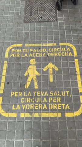 This video provides a static, overhead view of a gray tiled sidewalk in L'hospitalet De Llobregat, Spain, under daylight conditions. The main subject is a large, rectangular yellow sign painted directly onto the pavement. The yellow paint appears somewhat faded and chipped in various spots, indicating wear from foot traffic and exposure.

Inside the yellow border of the sign, two lines of text are prominently displayed, along with two yellow pictograms. The top line of text reads in Spanish: "POR TU SALUD, CIRCULA POR LA ACERA DERECHA," which translates to "For your health, walk on the right sidewalk." Below this, a stylized figure of a person walking is depicted next to an upward-pointing arrow, suggesting forward movement and direction. The second line of text, written in Catalan, states: "PER LA TEVA SALUT, CIRCULA PER LA VORERA DRETA," also meaning "For your health, walk on the right sidewalk."

At the top of the frame, a square metal manhole cover or grate is visible, embedded within the sidewalk tiles. A few small pieces of debris, possibly cigarette butts, are scattered on the pavement near the grate. No people or specific activities are observed in the video; it is a still shot capturing the sidewalk and its directive signage.