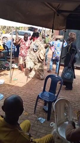 The video shows a gathering of people in an outdoor setting, possibly a backyard or event space, under an awning providing shade. The ground is paved with red bricks.

A central figure in the scene is a person dressed in a full-body costume made of straw-like material, with a mask covering their face. They are accompanied by at least two other men; one wears a red and white patterned shirt and khaki shorts, and the other is dressed in a green shirt. These men appear to be interacting with the costumed figure, possibly guiding or assisting them.

Several people are seated at tables, and others are standing or walking around. There are also chairs scattered around. A car is visible parked in the background, further suggesting this may be a residential setting.

The overall atmosphere appears festive and communal, hinting at a celebration or cultural event.