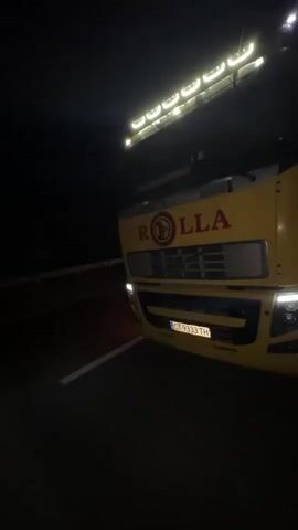 A yellow truck with multiple roof lights illuminated is seen at night. The camera begins facing the front of the truck, displaying "ROLLA" in red text on its grille and a license plate with "CT 7333 TH". The camera then pans to the right, revealing the side of the truck with "VEGETARI" text in red and yellow. The truck's headlights and side marker lights are active. A red light from another vehicle is visible in the distant background. The truck appears to be on a paved surface with a white line marking.