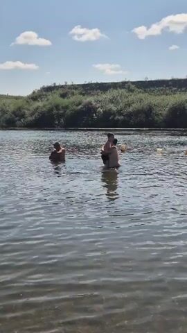 The video shows two people swimming in a river. The person on the left is further from the camera and appears to be standing with their head and shoulders above the water. The person on the right is closer to the camera and is embracing another person. They are both in the water, with their bodies mostly submerged. The river is wide and calm, with a shoreline covered in green vegetation in the background. The sky is blue with some white clouds. There are yellow buoys visible in the water to the right of the two swimmers. The scene appears to be a recreational activity on a sunny day.