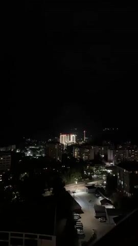 A nighttime aerial shot of a city with buildings and streetlights. In the distance, there is a large illuminated building. A thunderstorm rolls in, indicated by flashes of lightning illuminating the dark, ominous clouds over the mountains in the background.