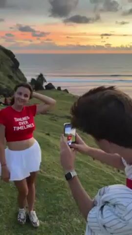 A young woman is posing for a photograph on a grassy hillside overlooking the ocean. She is wearing a red crop top with white lettering, a white miniskirt, and sneakers. A young man is taking her picture with a smartphone. He is wearing a striped shirt. The sky is colorful with the setting sun. There are other people on the hillside in the background.