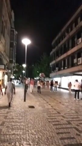 A nighttime street scene with people walking along a cobblestone sidewalk. A lamp post illuminates the path. Buildings line both sides of the street, some with shop windows. A group of people walk together in the center of the frame. The scene is dark with artificial lighting.