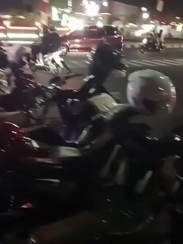 This video captures a nighttime scene on a paved walkway next to a street. Several motorcycles are parked on the walkway. In the background, vehicles are driving on the street, including a red van. The area is illuminated by streetlights and decorative lights, some of which cast colorful glows on the trees. Two red and white striped poles are visible, possibly marking an entrance or a boundary. The overall atmosphere suggests an urban or commercial area at night.