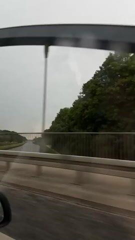 The video appears to be shot from inside a vehicle, looking out the window. The view is of a bridge over a body of water, possibly a canal. There are trees and greenery on either side of the waterway, and the sky is overcast. The camera is moving, suggesting the vehicle is in motion. The interior of the car is partially visible, including a portion of the dashboard and a side mirror. The overall impression is of traveling through a somewhat dreary, possibly rainy, landscape.