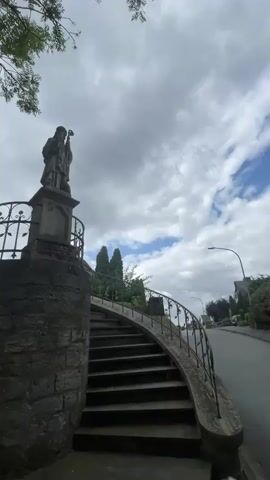 The video shows a view of a stone statue of a person holding a staff, standing on a stone pedestal. The pedestal is on top of a round stone structure. A curved metal railing leads up a set of stone stairs to the right of the pedestal. In the background, a large building with a pointed roof and spire can be seen, which appears to be a church. Trees and foliage are visible in the foreground and background. The sky is overcast with clouds. The camera pans slightly to the left, revealing more of the church and the surrounding greenery.