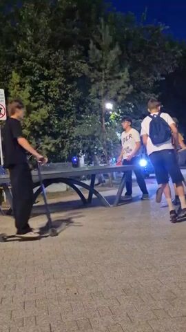 The video shows a nighttime outdoor scene. A group of people are near a ping pong table. In the foreground, a person on a scooter rides past the table. In the middle ground, a person in a white t-shirt stands by the table, holding a paddle. Another person in a black t-shirt walks away from the table. In the background, a vehicle with lights is visible, and more people are standing around. There are trees and streetlights in the scene. A sign with a dog symbol is on a pole to the left. The ground is paved with interlocking stones.
