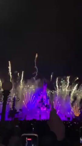 The video shows a nighttime fireworks display at Disneyland, with Sleeping Beauty's castle as the focal point. The castle is illuminated with purple and blue lights, while fireworks explode in the sky, producing colors like gold, white, and a fiery orange. The fireworks create a spectacular display that surrounds the castle, and the crowd in the foreground is seen holding up their phones to record the event. A street lamp is visible on the left side of the frame.
