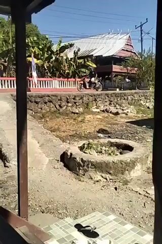 The video shows an outdoor scene on a sunny day. In the foreground, there are two pairs of shoes placed on a tiled surface. One pair is black flip-flops, and the other pair appears to be white clogs. Behind the shoes, a dry, rocky area is visible with a circular concrete structure in the middle, containing some green plants. Further back, a stone wall borders a raised area with a red and white fence. Beyond the fence, a traditional-looking house with a steep, metallic roof is situated amongst lush green trees. To the left of the house, a person is seen riding a motorcycle on a paved road. Overhead, power lines are visible against a clear blue sky. The overall environment suggests a rural or semi-rural setting, possibly in a tropical or subtropical region.