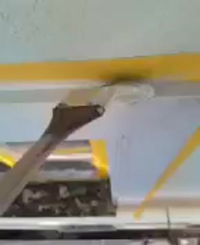 Here is a description of the video.
The video shows a hand holding a paintbrush and applying white paint to a surface. The surface appears to be a wall or ceiling, with yellow tape delineating the area to be painted. There are other items in the background, including what looks like a metal duct covered in yellow tape. The setting seems to be indoors.