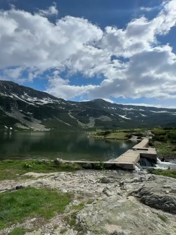 The video shows a serene mountain lake under a partly cloudy sky. In the foreground, a rocky, grassy area transitions to a concrete structure built across a small stream flowing out of the lake. This structure appears to be a small dam or spillway. The lake itself is calm and reflects the surrounding mountains. The mountains are steep and rugged, with patches of snow visible on their upper slopes, indicating a high altitude or recent snowfall. The sky is a vibrant blue with scattered, fluffy white clouds that cast shadows on the landscape. The overall atmosphere is peaceful and natural.