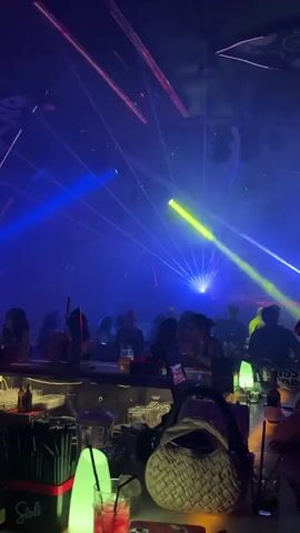 This media file depicts a vibrant nightlife scene, likely in a club or bar, in Khan Daun Penh, Cambodia. The primary activity is a party with a DJ, indicated by the pulsating laser lights and music, which create a dynamic and energetic atmosphere.

The scene is set inside a dimly lit venue adorned with an array of colorful laser lights, predominantly blue, pink, and green, which crisscross the space and illuminate the crowd. The lighting suggests it is nighttime.

In the foreground, a bar counter is visible, laden with glasses, bottles, and bar tools, including straws and mixers. A woven basket and illuminated table lamps add to the ambiance. Several people are seen at the bar, some appearing to be patrons and others possibly staff.

Beyond the bar, a crowd of people are gathered, silhouetted against the light show. They are engaged in socializing and enjoying the music and atmosphere. The overall mood appears festive and lively.

Visible text includes the brand name "Stoli" on black coasters or mats placed on the bar, and possibly on a dispenser. The image conveys a sense of excitement and social gathering, characteristic of a night out.