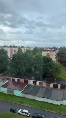The video captures a view from a window overlooking an outdoor scene. In the foreground, a cat is resting on a shelf. The window is partially visible, with a handle and some decorations. Outside, a row of garages with various colored roofs is visible, some appearing wet. Beyond the garages, there are trees and apartment buildings. The sky is overcast with clouds, suggesting a gloomy or rainy day. The road in front of the garages has a few parked cars and a motorcycle. The video pans slightly, shifting the perspective from the cat to the outdoor view.