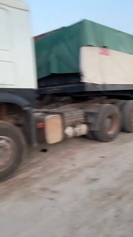 The video shows a dusty industrial area. It starts with a shot of a large truck with a green and white tarp covering its load. The camera then pans to the right, revealing a wide, dirt road and a person in an orange shirt walking in the distance. In the background, there's a long white wall, possibly surrounding a factory or industrial complex. Silos and a crane are visible behind the wall. The sky is a light blue, suggesting it's either early morning or late afternoon. The video seems to be taken from a moving vehicle.
