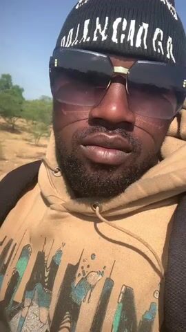 The video shows a close-up, selfie-style shot of a Black man wearing a black beanie with white text and large, dark-lensed sunglasses. He has a beard and is wearing a tan hoodie with a graphic design. A black backpack is visible over his shoulders. The background appears to be a dry, outdoor environment with sparse vegetation and a clear blue sky. The lighting suggests it is daytime, likely sunny. The man's expression is neutral. The text on the beanie is partially visible and appears to be a logo or brand name, possibly "BALENCIAGA". The overall impression is that the person is outdoors, possibly traveling or exploring. The scene is set in Gobbiya, Nigeria.