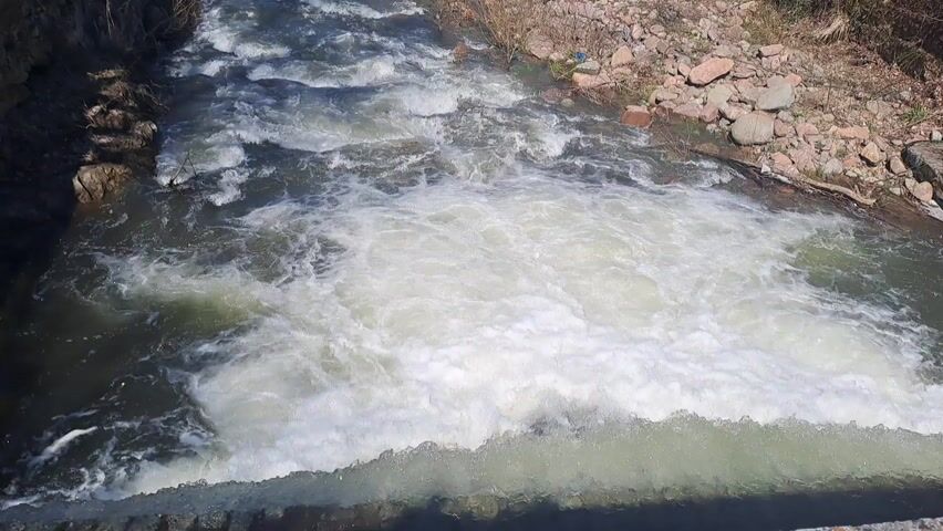 The video shows a river flowing through a narrow channel. The water is turbulent and foamy, creating white water as it flows downhill. The banks of the river are lined with rocks, sparse vegetation, and some sections are reinforced with stone walls. Beyond the riverbanks, a park-like setting with grass and trees is visible. The river is in the foreground, and the background is blurred. There are no people in the video.