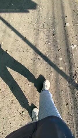 The video shows a first-person perspective of a person walking on a dirt path. The person is wearing light-colored athletic shoes, light blue pants, and a dark jacket. Long shadows are cast on the path, indicating it is a sunny day. The ground is dry and dusty, with some scattered debris and vegetation visible along the edges. The person's legs and feet are visible as they walk, and the camera follows their movement. The scene appears to be outdoors, possibly in a rural or unpaved urban area.