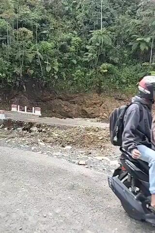 A group of motorcyclists navigate a damaged and uneven road. In the foreground, a lone motorcyclist rides on the right side of the road, moving away from the camera. Behind him, a cluster of three other motorcycles approaches, also traveling in the same direction. The road surface is a mixture of gravel and dirt, with visible damage and debris. To the left of the road, a steep, vegetated hillside rises. To the right, a section of the road appears to have collapsed, with a steep drop-off and piles of dirt and rocks. A red-railed bridge is visible in the middle ground, with some of the motorcyclists crossing it. A signpost with text is present near the bridge entrance. The sky is overcast.