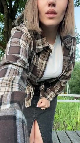 A young woman poses outdoors in daylight, under a large tree with a white fence behind her. She is wearing a long dark denim skirt with a slit, a white t-shirt, and a brown and beige plaid shirt tied at the waist.  She initially arranges her hair, looks at the camera, then puts her hand in her pocket and turns her head. The ground is covered with greenery and a wooden boardwalk.