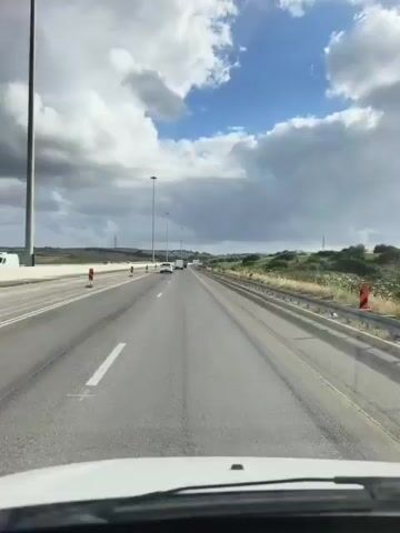 A vehicle is driving on a highway. The road is mostly straight, with some roadwork cones on the left side. Other cars are visible in the distance. The sky is a mix of blue and white clouds. There is green vegetation on the side of the road. The view is from inside the vehicle, looking out through the windshield.