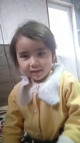 A young girl with dark hair and eyes, wearing a yellow sweater with a white faux fur collar, is the central figure in the video. She looks directly at the camera, smiles, and says "Assalomu alaykum". She holds a thin, tan-colored object in her hands, possibly a bow. The background includes a white wall with grey veining, a partial view of a light-colored door, and a dark vertical structure with wires visible. The video seems to be taken indoors in a domestic setting.