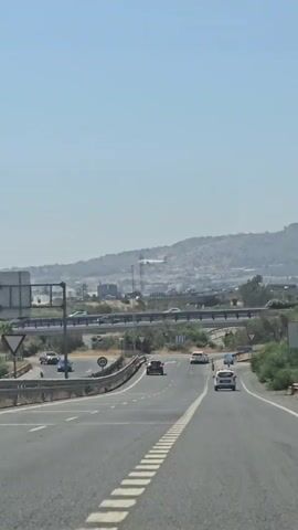 The video shows a view from a vehicle moving along a highway on a clear, sunny day. The highway has multiple lanes and is bordered by a guardrail. Several cars are visible on the highway, moving in the same direction as the camera. In the background, there are buildings, trees, and a distant hill or mountain range. A speed limit sign indicating 40 is visible on the side of the road. A train track runs parallel to the highway in the middle distance, with a train appearing to be in motion on it.