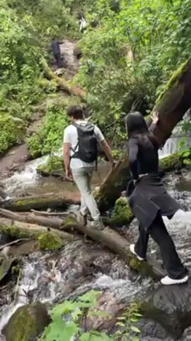 A low-angle shot shows three people carefully navigating a flowing stream. A man with a backpack is in the lead, stepping across fallen logs. A woman in a black outfit follows, holding onto another log for balance. A third person, wearing a red cap, is behind her. The stream is surrounded by lush green vegetation, and other people are ascending a steep path on the hillside in the background.