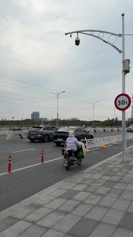 The video shows a street scene on a cloudy day. Cars drive by on a two-lane road, separated by white and yellow barriers. The sidewalk is tiled in a grey and white pattern, and a sign indicates a weight limit of 30 tons. Power lines stretch across the sky.  A security camera is mounted on a lamppost. People on scooters drive along the sidewalk. In the background is a modern building.