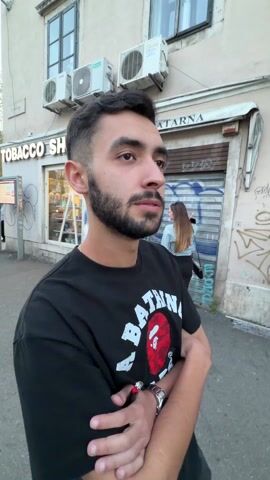 A young man stands in front of a Tobacco Shop on a city street. He has dark hair, a beard, and is wearing a black t-shirt with a red and white logo that says "BATHING APE" on it. His arms are crossed. Behind him are people walking on the sidewalk, and there is a closed store with graffiti on the metal shutter. There are several air conditioning units on the building above the store. A streetlight is visible on the left.