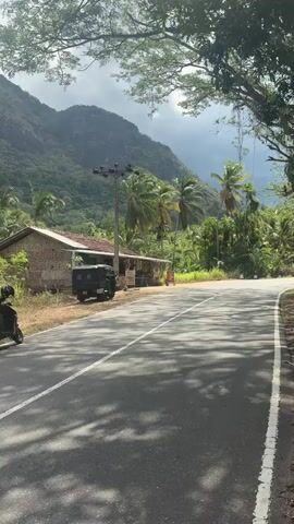 A motor scooter is parked on the left side of a paved road. Beside the scooter is a small, brick building with a corrugated metal roof. A dark-colored auto-rickshaw is parked in front of the building. The road curves to the right. Lush green trees and palm trees are visible on the right side of the road. In the background, a large, lush green mountain looms under a cloudy sky. A large, red bus drives down the road from left to right, passing the building and auto-rickshaw. The bus has multiple windows, and people can be seen inside. Sunlight filters through the trees, casting shadows on the road.