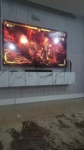 The video shows a widescreen television mounted on a wall. The screen displays a video game battle with a red and fiery background. Two figures are engaged in combat, performing martial arts moves. Below the TV is a white shelf unit with a marble pattern. A PlayStation 5 gaming console is visible to the right of the television. The room has textured, gray striped wall panels and a patterned rug on the floor.