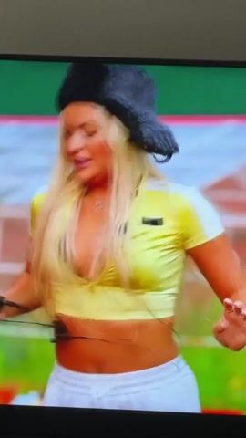 A blond woman in a yellow crop top and white pants wears a black fur hat. She dances with a man in a maroon shirt and white pants outdoors in front of a wooden cabin. The woman holds a black object in her left hand. She gestures with her right hand. The man holds her left hand and pulls her slightly. A pink tablecloth covers a table to her left. The ground is covered in green grass and some white objects.
