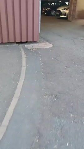 The video shows a view from a person's perspective walking into a covered parking garage. Cars are parked in rows on either side of the garage. The floor is asphalt and appears to be cracked in places. There is a metal fence and a brown wall on the left side of the frame. The lighting in the garage is dim.
