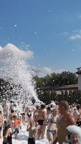 A crowd of people are enjoying a foam party outdoors on a sunny day with a clear blue sky. A large amount of white foam is being sprayed into the air from the left side of the frame, creating a cloud of foam that is falling onto the people. The people are dressed in swimwear and appear to be having fun, with some raising their hands in the air. The background shows green trees and some buildings.