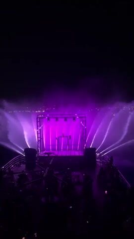 This is a vertical video of a nighttime outdoor stage performance. The stage is a raised platform with a rectangular metal frame above it, equipped with several lights. There are water fountains spraying upwards and outwards from behind the stage, illuminated by colored lights. The lights on the stage and fountains change colors throughout the video, cycling through purple, yellow, and green. Silhouettes of an audience can be seen in the foreground, looking towards the stage. In the background, distant city lights are visible on a horizon. The overall atmosphere is dark and dramatic, with a focus on the light and water spectacle.