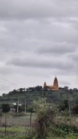 A vertical, outdoor, daylight, medium shot shows a golden Hindu temple situated atop a lush green hill. The sky is overcast with gray clouds. In the foreground, a dirt patch is covered with dry grass and a chain-link fence. A person, whose back is to the camera, walks away from the viewer towards the fence. The person is wearing a white shirt and dark pants. The camera slowly zooms in on the temple.