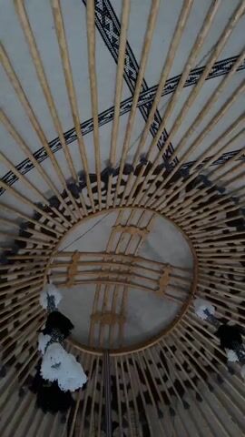 The video shows the interior of a yurt, a traditional nomadic dwelling. The perspective is looking upwards towards the roof structure. The roof is made of wooden poles radiating from a central circular frame, called a "tuno." Decorative fabric with geometric patterns lines the interior walls and ceiling of the yurt. Several tassels, white and black, hang from the roof structure. The lighting is dim, typical of the interior of a felt-covered structure.