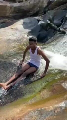 A young man is filmed sliding down a smooth, rocky, wet area that empties into a pool of water. The rocky area appears to be a natural water slide. The man is wearing a white tank top and dark-colored shorts. He is sitting and bracing himself with his hands as he slides down into the water. The water is clear, and the rocks around the area are various shades of gray and brown.