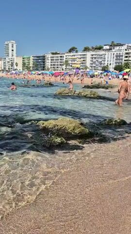 The video shows a crowded beach on a sunny day. People are swimming in the clear blue water, sunbathing on the sand, and relaxing under colorful umbrellas. In the background, there are several buildings, likely hotels or apartments, lining the coast. The water is calm with gentle waves lapping the shore, and there are some rocks visible in the shallow areas. The overall atmosphere appears to be one of leisure and recreation.
