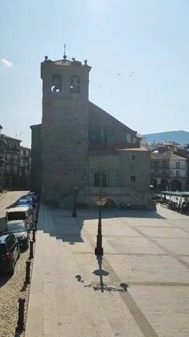 The video shows a view of a town square with a stone church on the right and a street lined with parked cars on the left. The sky is bright and clear, suggesting it is daytime. A lamppost with red flowers in a planter stands in the middle of the square. The architecture of the church is visible, including its bell tower and arched windows. The buildings on the left appear to be residential or commercial. Some birds are seen flying in the sky. The ground of the square is paved with stone tiles, and a patterned walkway leads towards the church. The overall scene is quiet and appears to be a sunny day in a European town.