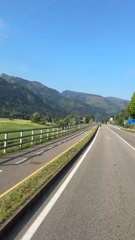 The video shows a view from a moving vehicle on a road. The road has a dedicated bike lane to the left, separated by a yellow line and grass. The bike lane is marked with white dashed lines and a bicycle symbol. A white metal fence runs alongside the bike lane. Beyond the fence is a green grassy field. In the background, there are mountains covered in trees. The sky is clear and blue. There are road signs visible, including a blue circular sign with a bicycle, a rectangular sign with "36", a blue circular sign with a bicycle and a red diagonal line, a pedestrian crossing sign, and a yellow rectangular sign with "SKI BUS" and a bus symbol. There is also a bus stop shelter with a bench. A person is walking on the sidewalk near the bus stop. The road continues into the distance, with a few other vehicles visible.