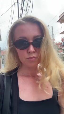 A woman with blonde hair and sunglasses walks down a street, filming herself. She is wearing a black top and a black jacket. The street is lined with buildings and has electrical wires overhead. There are several motorcycles in the background.