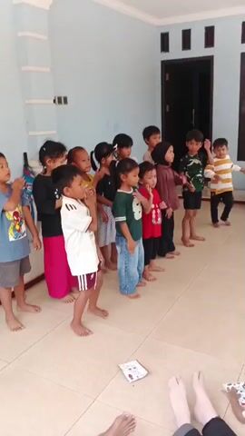 A group of children stand in a line facing forward. They are performing a series of synchronized hand movements, including bringing their hands to their chests, clapping, and pressing their hands together as if in prayer. The children are barefoot and dressed in casual clothing. The background is a light blue wall with a white pillar and a dark doorway. A few scattered papers lie on the tiled floor. The children appear to be engaged in an activity, possibly a song or a dance.